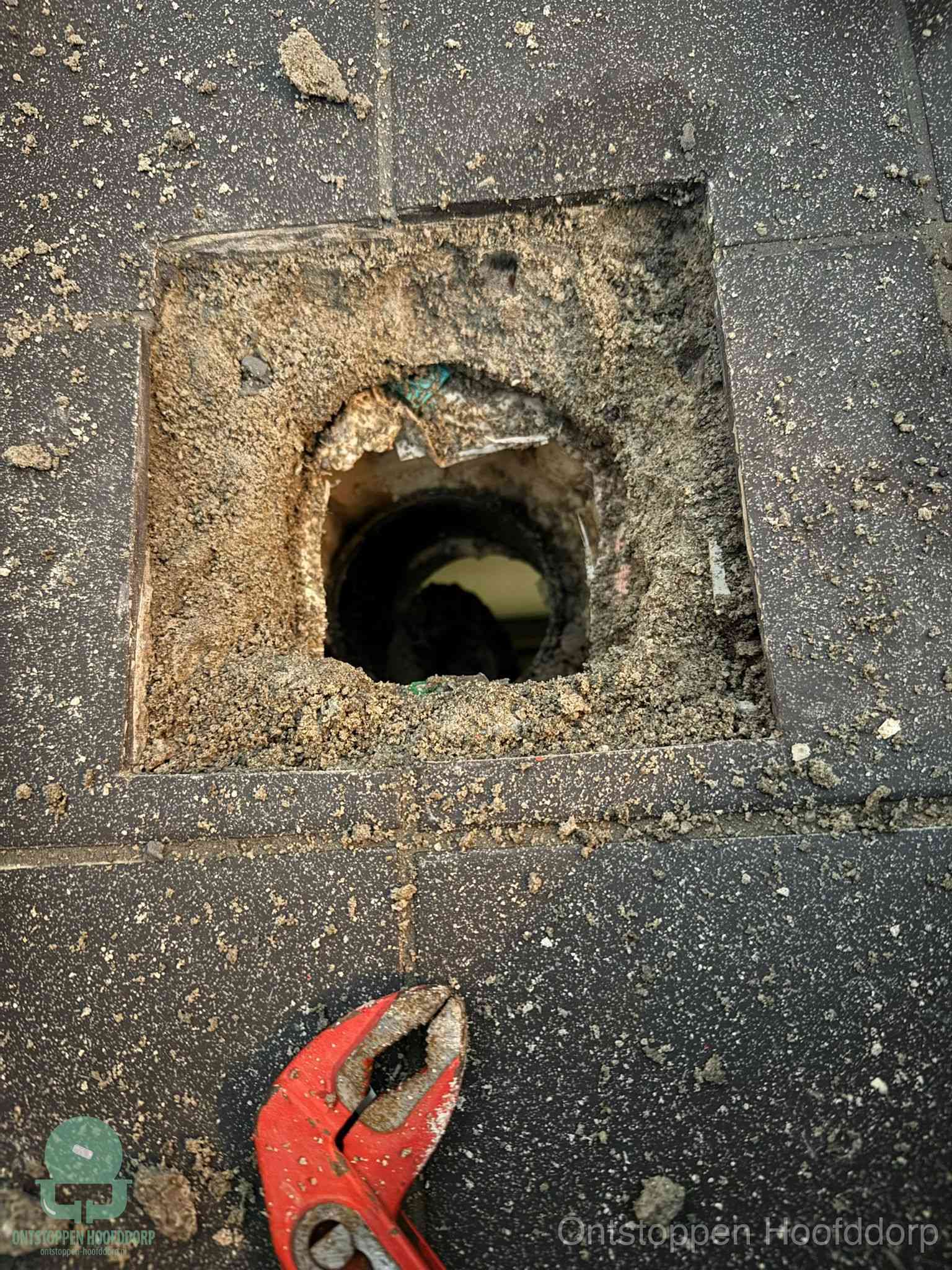 Een geopend rioolputje in een betontegelvloer met losgemaakt rooster en een zichtbare ronde afvoerbuis, het achterhalen waar de afvoerverstopping zit in Hoofddorp