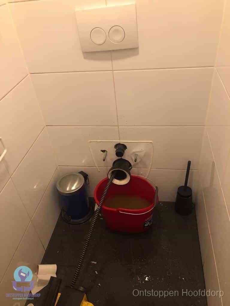 Een ontstopte wand-wc met verwijderde toiletpot waar een ontstoppingsveer in de wanduitgang verdwijnt, het achterhalen waar de verstopping zit in Hoofddorp