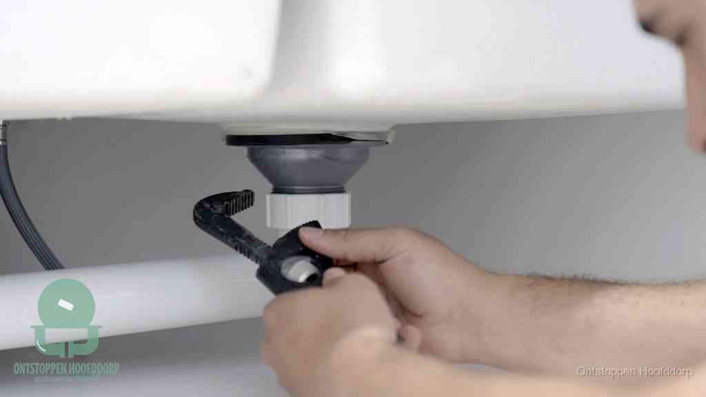 Twee handen draaien met een waterpomptang de wartelmoer van een kunststof sifon onder een wastafel los om de sifon schoon te maken in Hoofddorp
